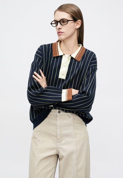 Μπλε σκούρο πλεκτό πόλο με ριγέ μοτίβο, καφέ ριμπ λαιμόκοψη, ανοιχτό μπεζ κουμπιά και μπεζ παντελόνι κορντόρι με ίσια γραμμή και απλή υφή.