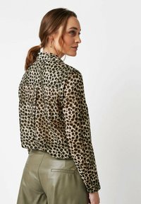 Woman with brown hair in a ponytail wearing gold hoop earrings, a long-sleeve leopard print top, and olive green pants, facing sideways.