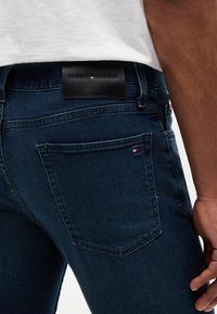 Close-up af en person iført mørkeblå jeans med en baglomme og et sort mærke med Tommy Hilfiger-logoet.