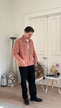 Jeune homme portant une veste matelassée saumon et un pantalon noir, debout dans une pièce moderne avec des lanternes décoratives, une chaise avec de la fourrure et une table d'appoint décorée.