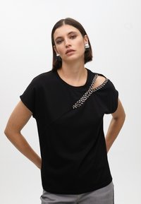 Black short-sleeve top with asymmetrical neckline, featuring a decorative band of clear gemstones across the shoulder. Smooth texture.
