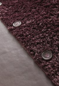 Lilla tekstureret stof med en fuzzy overflade præsenterer to runde knapper. Et glat, gråt materiale er synligt nedenunder.