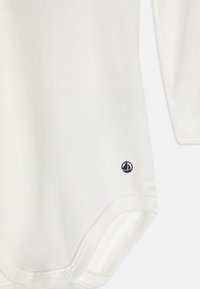 Weißer Langarm-Bodysuit aus weicher Baumwolle, mit Rundhalsausschnitt und kleinem Marine-Segelboot-Logo unten links.