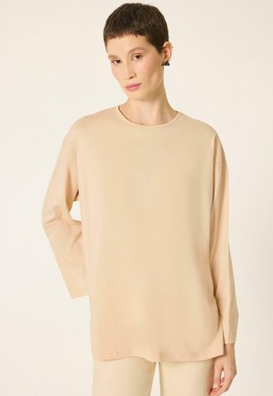 Femme aux cheveux courts portant un haut beige ample à manches longues et un pantalon de couleur claire, debout devant un fond clair uni.
