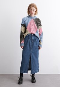 Mehrfarbiger Strickpullover mit einem Zopfmuster in Blau, Pink und Beige; Denim-Maxi-Rock mit einem Schlitz vorne; getragen mit schwarzen Schuhen.