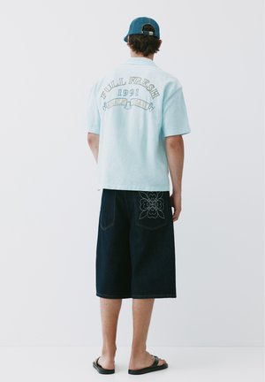 Homme portant une chemise bleu clair avec un texte au dos, un short foncé au genou avec un motif floral sur la poche, une casquette bleue et des sandales noires, debout de dos.