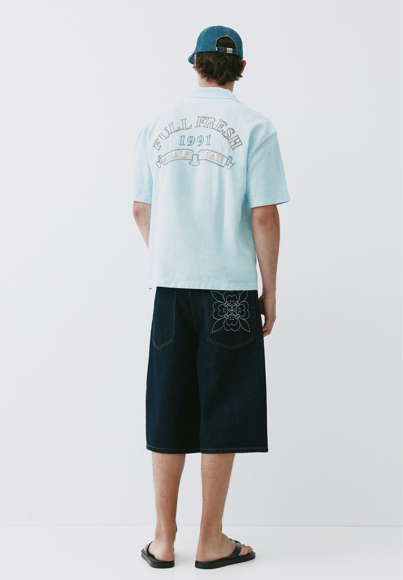 Homme portant une chemise bleu clair avec un texte au dos, un short foncé au genou avec un motif floral sur la poche, une casquette bleue et des sandales noires, debout de dos.