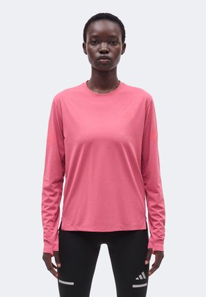 Žena koja nosi dugačku ružičastu majicu s okruglim izrezom i crne tajice s reflektirajućim Adidas logotipima na bijeloj pozadini.