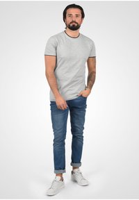Camiseta gris de manga corta con ribetes negros, combinada con jeans azules y zapatillas blancas. El modelo tiene tatuajes y está de pie con las manos en los bolsillos.