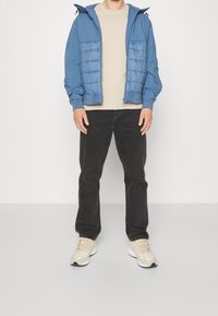 Muž v modré péřové bundě s kapucí, béžovém svetru, černých džínách a bílých teniskách, stojící na pozadí jednoduchého světlého pozadí.