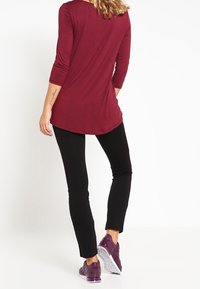 Burgundy långärmad topp med rundad nederkant, parat med svarta tajta byxor och lila träningsskor. Slätt tyg, enkel design och åtstramad passform.