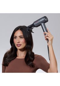 Femme aux cheveux ondulés et foncés séchant ses cheveux avec un sèche-cheveux Revlon gris muni d'un embout concentrateur sur un fond gris uni.