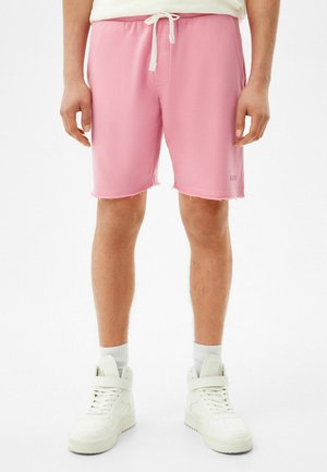Man draagt lichtroze shorts met trekkoorden, witte sportsokken en witte hoge sneakers, staand tegen een effen achtergrond.