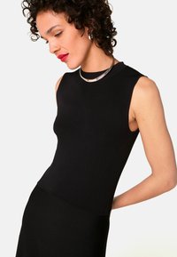 Schwarzes ärmelloses Top, taillierte Passform, gerippte Textur. Mit einer silbernen Halskette geschichtet, posiert das Model mit einer subtilen Biegung. Schlichte, elegante Styling.