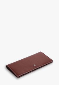 Cartera de cuero marrón con un diseño elegante, que presenta un pequeño emblema plateado de calavera y huesos cruzados. Superficie texturizada, forma rectangular.