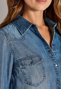 Denim overhemd met een lichtblauwe naar donkerblauwe gradient, puntige kraag, twee borstzakken met klep en zilveren drukknoopjes. Zachte textuur.