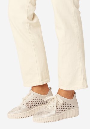 Voeten in crèmekleurige sneakers van gebreide stof met dikke zolen en witte broeken die boven de enkels korter zijn gesneden.