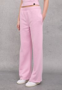 Pantalones de chándal rosa claro hechos de un tejido suave. Presentan un ajuste holgado, bolsillos laterales y un logo bordado en la parte frontal.