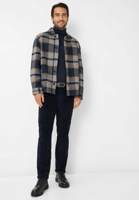 Gevlekt flanellen shirt in blauw, grijs en bruin, gecombineerd met een marineblauwe coltrui en donkere ribfluwelen broek. Zwarte veterschoenen maken de outfit compleet.