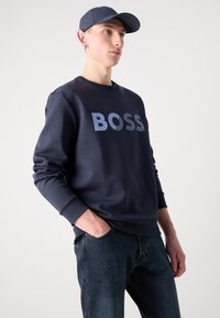 Μπλε ναυτικό φούτερ με λογότυπο "BOSS" σε πιο ανοιχτό μπλε, με στρογγυλή λαιμόκοψη και μακριά μανίκια. Συνδυάζεται με σκούρα τζιν και καπέλο σε ταιριαστό χρώμα.