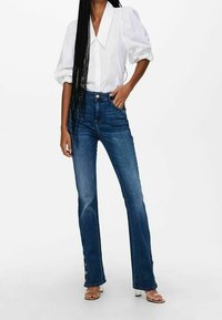 Femme portant un chemisier blanc à manches bouffantes, un jean bleu taille haute avec des boutons à l'ourlet, et des sandales à talons transparents.