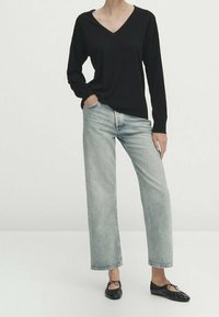 Personne portant un haut noir à manches longues avec col en V, un jean bleu clair à jambes droites, et des ballerines noires, debout avec une main dans la poche.