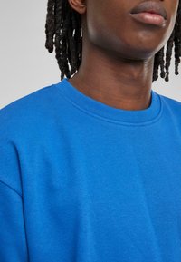 Blå bomullssweatshirt med rund hals, med en jevn tekstur og standard passform, som delvis viser bærerens hals og ansikt.