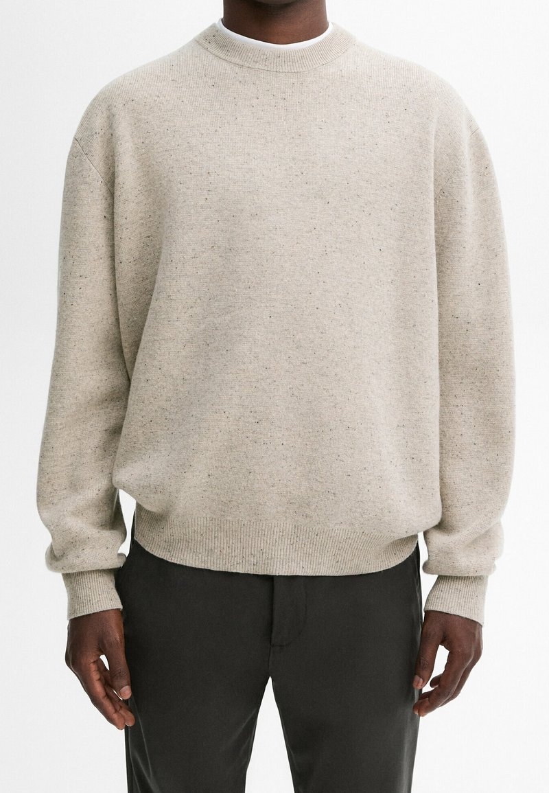 Personne portant un pull beige col rond avec une texture tachetée et un pantalon noir, debout sur un fond blanc uni.