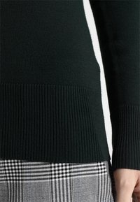 Pull tricot vert foncé avec ourlet et poignets côtelés, associé à une jupe à motif à carreaux noir et blanc. Texture lisse et coupe ajustée.