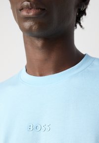 Κοντινό πλάνο ενός ατόμου που φοράει ένα ανοιχτό μπλε μπλουζάκι με στρογγυλή λαιμόκοψη, το οποίο έχει ανάγλυφο κείμενο "BOSS" στο στήθος και ένα μικρό σκουλαρίκι.