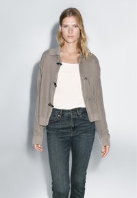Beige knit cardigan with a collar, featuring dark buttons, paired with a white top and dark jeans. Smooth texture and relaxed fit.