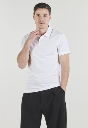 Uomo con polo bianca a maniche corte e pantaloni neri, che tocca il colletto con una mano, in piedi davanti a uno sfondo chiaro e uniforme.