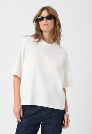 Top blanco de manga corta con cuello redondo, textura suave, corte relajado, combinado con jeans azul oscuro. Diseño simple y minimalista.