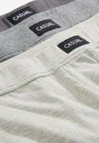 Tre paia di pantaloni corti casual in grigio chiaro, grigio scuro e bianco sporco. Ognuno presenta un'etichetta nera con la scritta "CASUAL." cucita sulla cintura.
