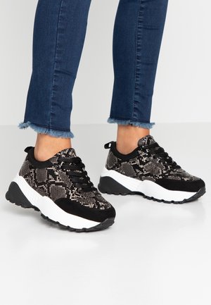 Pieds portant des baskets à motifs serpent noir et blanc, associées à un jean skinny bleu foncé à ourlet effiloché, sur un sol clair.