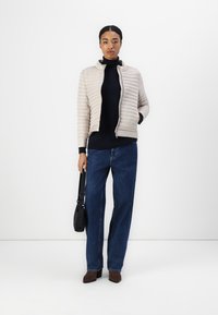Lyse beige quiltet jakke med ståkrage, glidelås, båret over en mørk høyhalset genser og blå vide jeans; svart skulderveske.