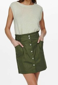 Hellgrünes ärmelloses Strickoberteil kombiniert mit einem hoch taillierten olivgrünen Knopfrock mit Seitentaschen und gerafftem Bund.