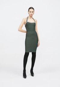 Πλεκτό φόρεμα σε σκούρο πράσινο χρώμα με εφαρμοστή σιλουέτα, λαιμόκοψη halter και χάντρες ως διακοσμητικά κατά μήκος του ντεκολτέ. Το μοντέλο φορά μαύρες καλσόν και τακούνια.