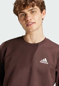 Brun bomullströja med rund hals och vit Adidas-logga på vänster bröst. Tyget har en mjuk och slät textur.