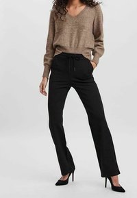 Pull en V tricoté marron avec texture côtelée associé à un pantalon noir à cordon de serrage. Le mannequin porte des chaussures à talons hauts noires.
