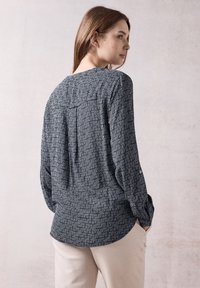 Camicia a maniche lunghe blu navy con un motivo geometrico bianco, caratterizzata da una spalla posteriore e polsini con bottoni, indossata con pantaloni chiari.