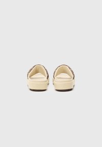 Brown textile slip-on shoes with a beige sole, featuring a textured upper and wide opening for easy wear. Minimalistic design with logo detail.