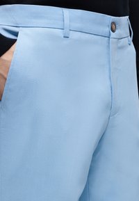 Ανοιχτό μπλε παντελόνι με κουμπί και θηλιές για ζώνη, φορεμένο με το χέρι στην τσέπη, σε μαύρο φόντο με πάνω μέρος ρούχου.