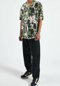 Korte mouwen, knoopshirt met een groen, paars en wit abstract patroon, gecombineerd met zwarte broek en multicolore sneakers.