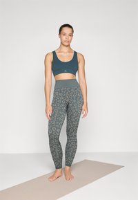 Mørk teal idretts-BH kombinert med leopardmønstrede leggings i nyanser av grått og teal; glatt tekstur, høyt liv og logodetalj på BH-en.
