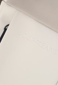 Borsa bianca con parte superiore in tessuto beige texturizzato, dotata di un logo "JOOP JEANS" in rilievo e di una tracolla nera in accentuazione. Materiale liscio e forma rettangolare.