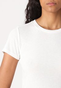 Camiseta blanca de manga corta hecha de algodón. Presenta un cuello redondo y una textura suave, con una forma ajustada que se adapta al cuerpo.