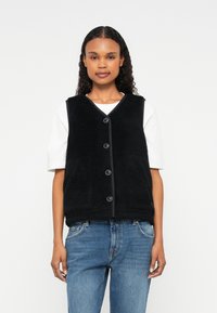 Black fleece vest with a V-neck, front button closure, and two pockets, worn over a white T-shirt and paired with blue jeans.