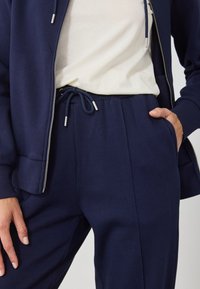 Sweatshirt azul-marinho com fecho e calças com cordão, apresentando punhos canelados e uma textura lisa e suave. Usado com uma t-shirt de cor clara.