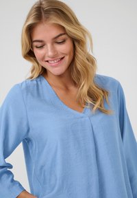 Blouse bleu clair à manches longues avec un décolleté en V, fabriquée dans un tissu doux et texturé et offrant une coupe décontractée. Le tissu semble léger.